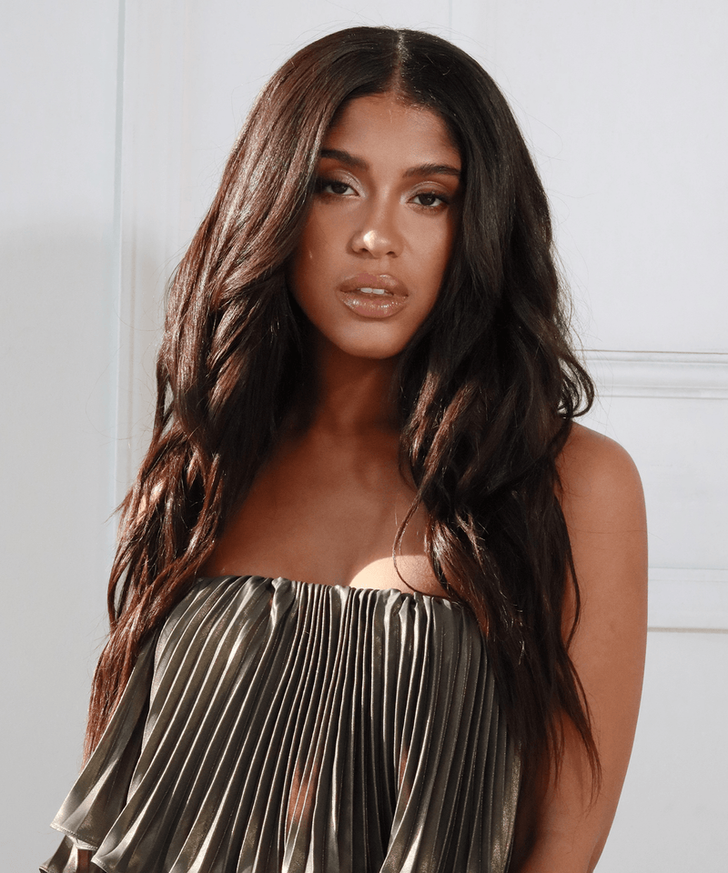 A woman with long, wavy dark hair enhanced by Perfect Locks extensions poses calmly in front of a white wall, exuding confidence.