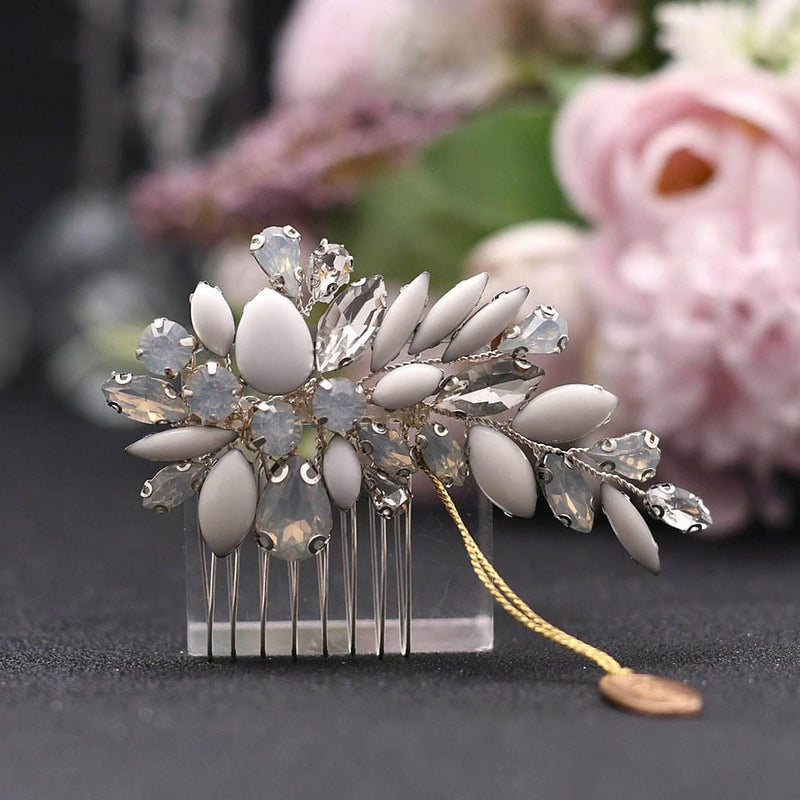A decorative hair comb with white and translucent gemstones in a floral design, gold chain and charm, on a blurred pink floral background.