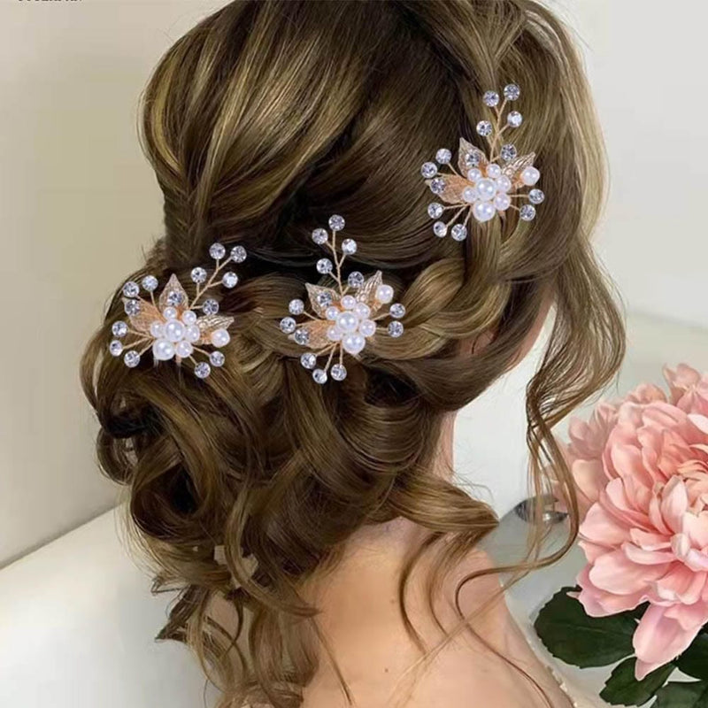 A woman with wavy brown hair in an elegant updo, adorned with pearl and crystal hairpins, sits near lush light pink flowers.