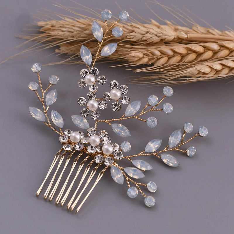 A decorative hair comb with gold wire, pearl-like beads, clear gemstones, and opal stones rests by wheat stalks on a gray surface.