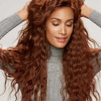 A woman with long, curly auburn hair in a gray turtleneck sweater looks down with a gentle smile, adjusting her hair gracefully.