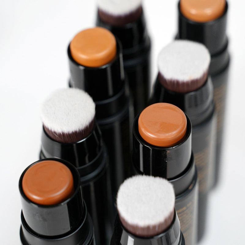 Sleek black makeup sticks with brown cream tops and soft white round applicator brushes arranged closely on a white background.