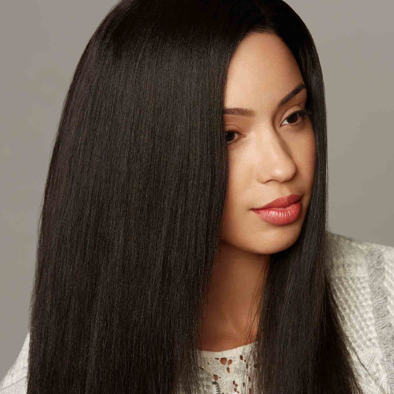 A woman with long, straight, shiny black hair and natural makeup wears a white textured top, gazing thoughtfully to the side against a plain background.