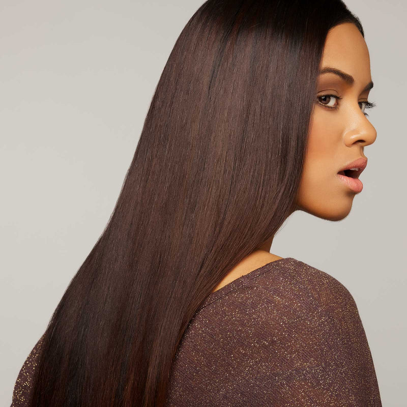 A woman with long, shiny brown hair glances over her shoulder in a sparkly brown top, her expression neutral against a plain light background.