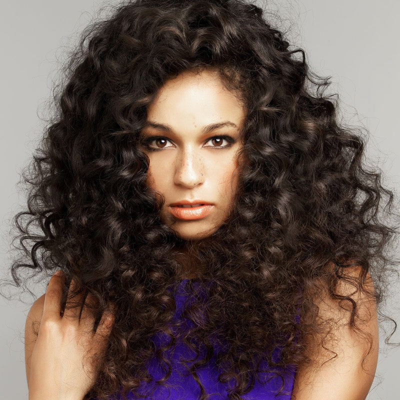 A woman with long, voluminous, curly dark brown hair and light skin wears a purple top, looking at the camera on a plain background.