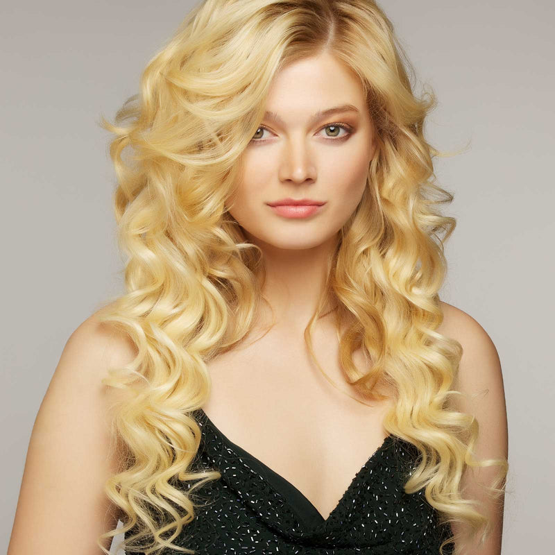 A confident woman with long, wavy blonde hair and fair skin wears a sleeveless black top, posing against a neutral backdrop.