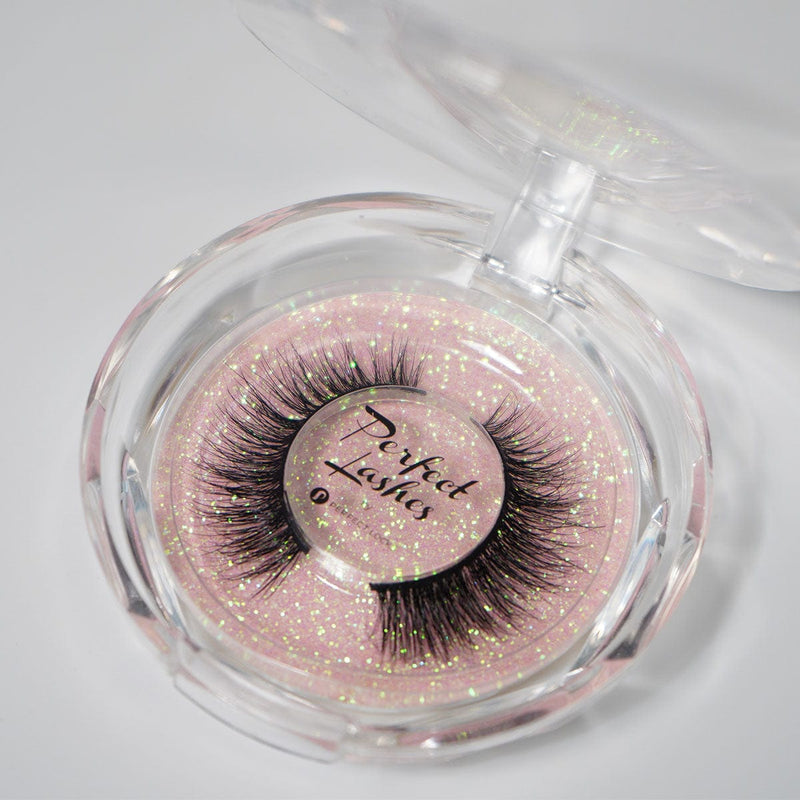 A round clear plastic case holds thick black vegan false eyelashes on a sparkly pink background labeled Perfect Lashes in the center.